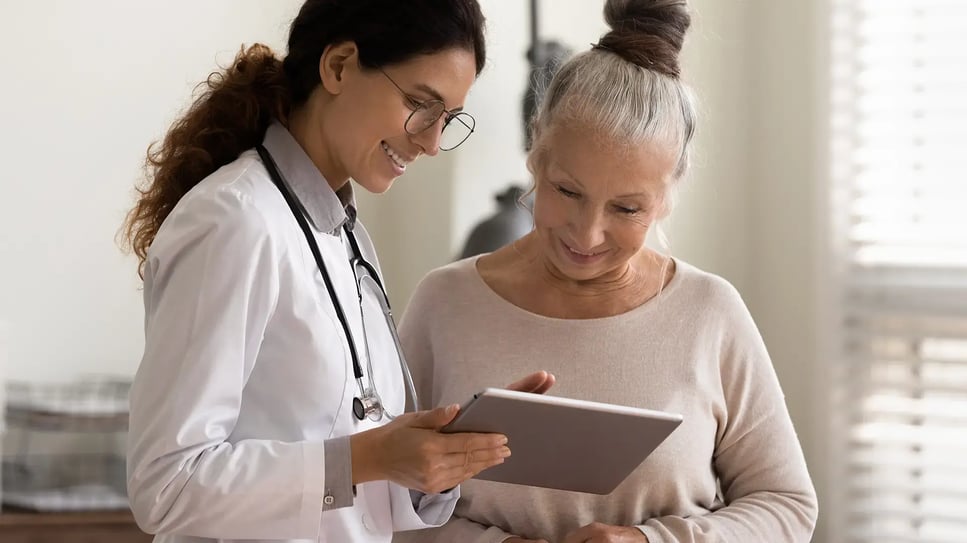 Happy female doctor and senior patient look at tablet screen discuss treatment or therapy on gadget. Smiling woman nurse and mature client use pad device discussing results in modern hospital
